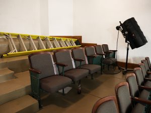 A section of theater balcony seating featuring vintage wood and fabric chairs, a yellow extension ladder resting on the stairs, and a large stage light on a stand.