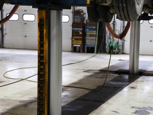 A heavy-duty vehicle lift in a garage bay, featuring a vertical chrome piston and a yellow safety locking rack.