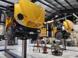 Two yellow school buses are raised on hydraulic lifts inside a large maintenance bay with their hoods open for mechanical repairs.