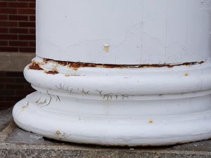 Close-up of a white painted column base with peeling paint and visible rust damage.