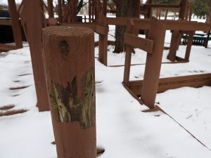 A close-up of a brown-painted wooden post on a snow-covered playground, showing significant peeling paint and weathered wood underneath.