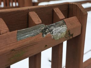 A close-up of a brown wooden railing on a playground where a large section of paint has chipped away, exposing grey, weathered, and splintering wood.