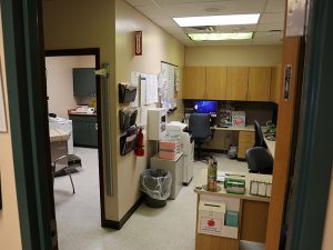 A cramped health office workstation area with desks, computers, and cabinets, and a doorway leading to a smaller examination room with a scale and medical equipment.