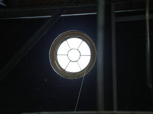 A small, circular window with a wheel-spoke muntin design set into a dark attic.
