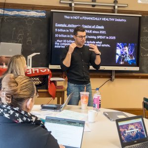 A teacher speaks in front of a screen that says "this is not a financially healthy industry".
