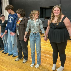 A group of performers stand side by side onstage holding hands, forming a line. Musical equipment and chairs fill the background behind them.