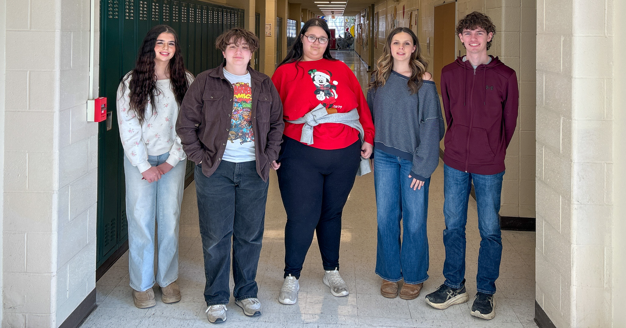 A group of five students standing side by side in a school hallway with lockers along the wall.