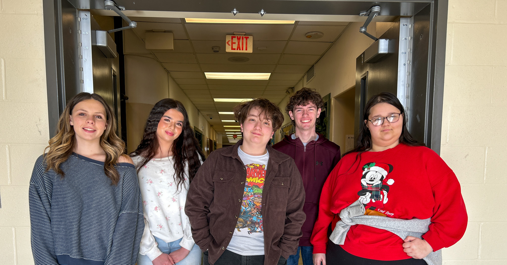 A group of five students standing together in a school hallway beneath a doorway with an exit sign overhead.