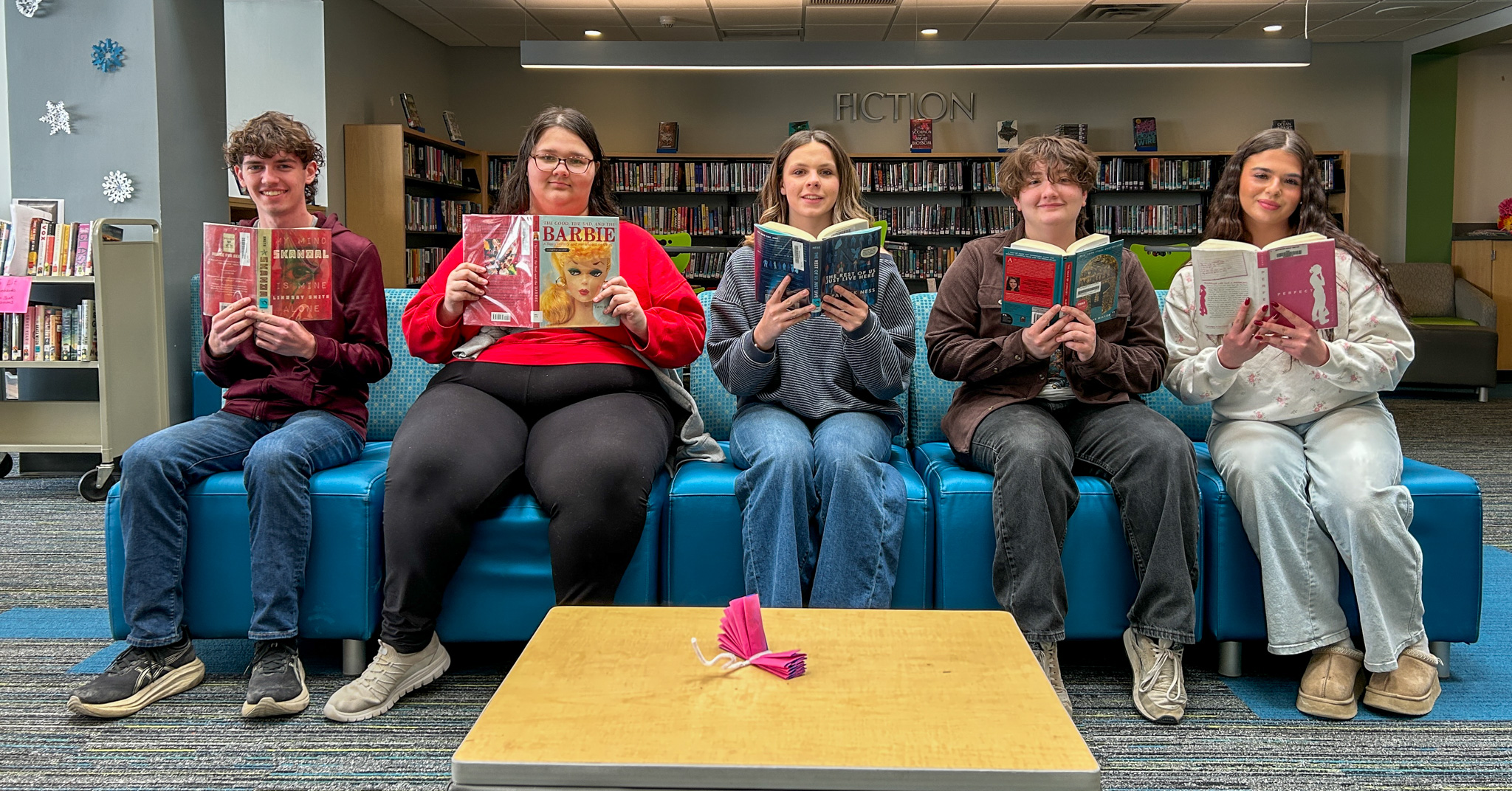 Five students sitting on a blue couch in a library, each holding an open book with shelves of fiction books behind them.