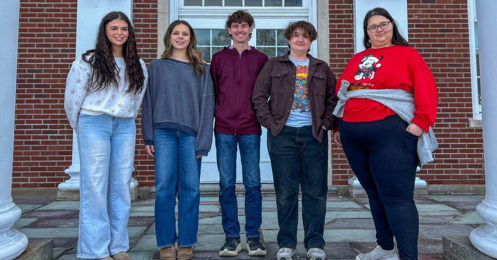 Five students standing in a row outside a brick school building with white columns.