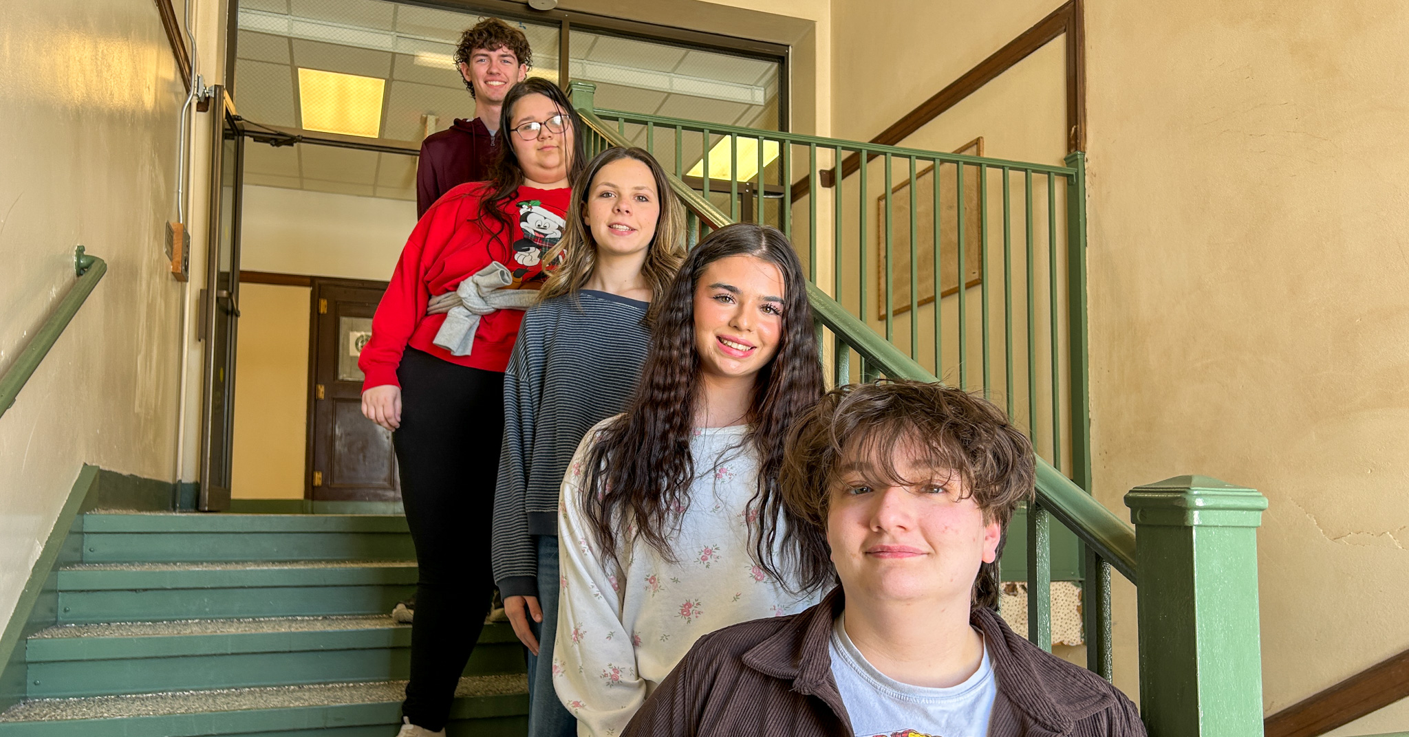 Five students standing on a staircase inside a school building, arranged in a staggered line down the steps.