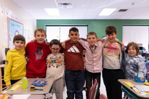 A group of elementary students stand together between classroom desks, posed closely with arms around each other. The room includes desks, chairs, and instructional materials, highlighting a school classroom setting.