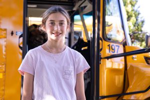 A student stands beside a yellow school bus with the bus door open. The side of the bus and identifying number are visible, emphasizing school transportation and daily operations.
