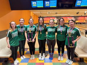 Members of the Middleburgh Knights girls bowling team stand together at a bowling alley wearing matching green team shirts. Several students wear medals and hold large trophies, with bowling lanes and pins visible behind them.