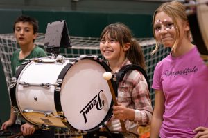 Students stand in a gymnasium at a school pep rally. One student holds a large bass drum while others stand nearby with face paint. A gym wall and netting are visible behind them, showing a school event setting.