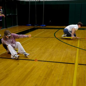 Two students wearing goggles move across a gym floor on low scooters during an activity marked with cones, with one student turning while the other moves in the opposite direction.
