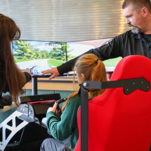 A student sits in a driving simulator with a red seat, while an adult points at a large screen showing a road scene and dashboard display, guiding the student during a safety demonstration.