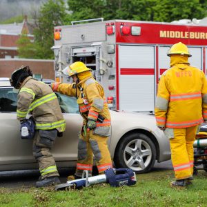 Firefighters wearing protective gear work beside a damaged car in front of a fire truck labeled “Middleburgh.” One responder leans into the vehicle while another stands nearby, and hydraulic rescue tools rest on the ground in the foreground.