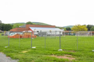 A chain-link fence encloses a grassy school field holding stacked playground or construction components, a white trailer, and soccer goals in front of a brick building.