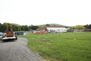 A steamroller sits on a flatbed trailer next to a fenced-in school field containing a modular trailer, a soccer goal, and wrapped construction materials.