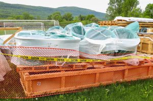 A construction staging area on a grassy field featuring large, circular, teal-colored components wrapped in clear plastic with caution tape surrounding.
