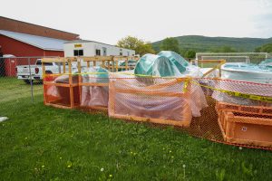 Wrapped construction materials and teal plastic slide segments sit on wooden pallets within an orange-fenced staging area on a school field, with a construction trailer in the background.