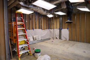 An A-frame ladder and construction lift stand in an unfinished room under bright overhead lights, featuring exposed wall framing, loose building materials, and protective white sheeting taped along the baseboards.
