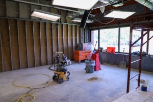An industrial shop vacuum sits on a dusty concrete floor inside a room under renovation, which features exposed wall studs, an orange tool box, a red fire extinguisher stand, and large windows looking outside.