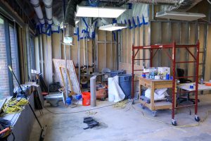 A room undergoing renovation is filled with construction equipment, featuring a red rolling scaffold, a wooden utility cart, and exposed framing walls prepped with blue painter's tape and plastic sheeting.
