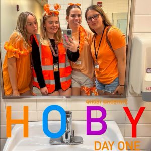 Four HOBY participants wearing orange-themed clothing pose for a mirror photo in a restroom on the first day of the seminar. The image includes the text "HOBY Day One."