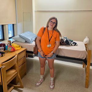 Cecilia stands in a college dorm room wearing an orange HOBY T-shirt, name badge and backpack. Two beds, a desk and personal belongings are visible in the room.