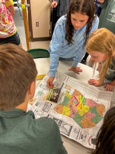 Students lean over a large map covered with handwritten notes while one student places a photograph on the map during a classroom activity exploring time capsule contents.
