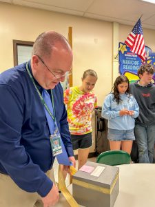 Superintendent Mark Place removes tape from a time capsule while members of the Class of 2038 watch from around a classroom table.

