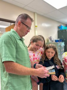 Teacher Terry Burton stands with students in a classroom as they examine photographs and other items removed from a time capsule.

