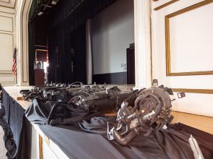 A row of black stage lighting fixtures rests along the front edge of an auditorium stage during a renovation project. The stage is framed by black curtains and decorative white-and-gold wall panels, with an American flag visible in the background. The lighting equipment appears staged for installation or replacement as part of auditorium upgrades.