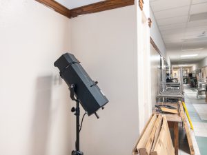 A stage lighting fixture mounted on a portable stand is positioned in a school hallway during an auditorium renovation project. Cardboard boxes and equipment are stacked nearby, while chairs are stored upside down on tables along the corridor in the background. The image highlights theater lighting equipment being prepared for installation or testing as part of ongoing auditorium upgrades.