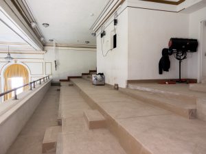 Upper-level seating area of an auditorium under renovation, showing stepped concrete risers, exposed wall-mounted equipment and cabling, and work materials placed along the aisle. Decorative white-and-gold architectural details and tall curtained windows are visible along the left side of the space.