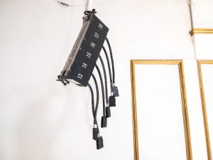 Close-up of a wall-mounted electrical distribution box labeled with numbers 13 through 18. Several thick black cables extend from the unit and terminate in hanging connectors against a white textured wall with gold trim.