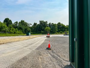 Road roller compacting gravel surface near building.