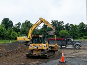 A construction site beside a transportation facility shows heavy equipment being used to improve field drainage. In the foreground, a yellow bulldozer sits on a dirt surface near an orange traffic cone. Behind it, a large yellow excavator loads soil into a black dump truck. Piles of excavated earth surround the equipment, with a tree line and overcast sky in the background. The work area has been cleared and graded in preparation for installation of a new drainage system.