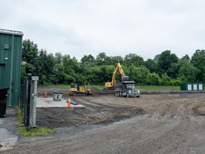 A wide view of drainage improvement work underway in a field adjacent to a transportation facility. An excavator transfers soil into a dump truck while a bulldozer operates nearby on a large, cleared construction area. The edge of a green maintenance building and a fenced utility area appear on the left. Freshly graded soil, construction materials and traffic cones are visible across the site, with dense trees lining the perimeter beneath a cloudy sky.