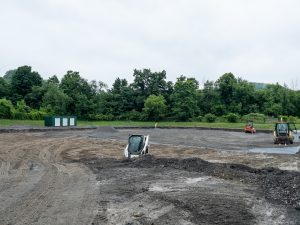 A large field beside a transportation facility is being regraded as part of a drainage improvement project. Construction equipment, including a skid-steer loader, a compact roller and a backhoe, is spread across the site. Workers are preparing a broad level area covered with gravel and soil, while mounds of aggregate and newly shaped ground indicate ongoing drainage installation. Trees surround the work zone under overcast conditions.