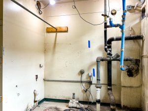 A utility room under renovation contains exposed plumbing, drain lines and electrical wiring. Several pipes run along the walls and floor, with wall openings and unfinished surfaces visible throughout the space as construction work continues.