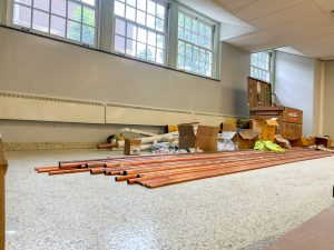 Construction materials, including copper piping, fittings and boxes, are staged inside a room with large windows. The floor and walls are finished, while tools and supplies indicate ongoing work as part of a renovation project.