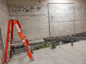 onstruction area inside a building shows an orange step ladder in front of a wall where finishes have been removed. A trench with exposed utilities and coiled green tubing runs along the base of the wall, with construction debris, a water bottle and utility connections visible on the concrete floor.