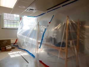 An interior room is sealed off for construction work with clear plastic sheeting taped from floor to ceiling. Sunlight streams through a large window, illuminating the temporary containment area and nearby construction materials.