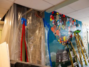 A hallway construction zone features clear plastic containment barriers and several ladders positioned near a colorful wall mural. The mural includes painted hot-air balloons and the text “Seniors 2010,” while renovation work continues around the protected area.