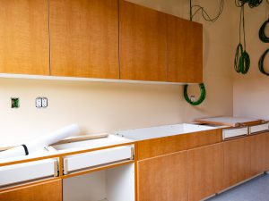 Wood-finish upper and lower cabinets are being installed in a new room. Several base cabinets and drawers remain unfinished, with countertops not yet in place. Coiled green wiring hangs from the wall near electrical outlets, indicating ongoing construction.