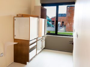 A partially completed room features newly installed wood-finish storage cabinets with several doors and drawers still open or awaiting installation. A large window overlooks a playground and school building outside. The room has finished flooring and painted walls, with construction work still underway.