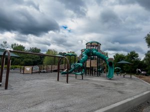 A newly constructed playground. The play area features a large tan and green play structure with multiple towers, enclosed tube slides, an open slide and climbing elements. In the foreground, the entire playground base is covered with a light gray crushed-stone surface that appears freshly installed and evenly graded. The stone material extends throughout the play zone, surrounding the equipment and creating a smooth, level foundation. A low concrete curb borders the edge of the playground, helping contain the loose aggregate surface. Brown metal swing supports stand over the gravel-covered ground, with swing seats not yet installed. Additional accessible play features, ramps and seating areas are visible in the background. Construction equipment can be seen near the edge of the site.